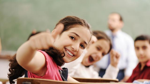 A child giving a thumbs up in a classroom with a teacher and other children.