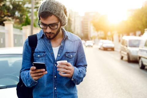 Young adult holding a cup walking in the street checking his phone