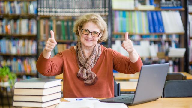 Older adult in a library with books and computer giving two thumbs up,