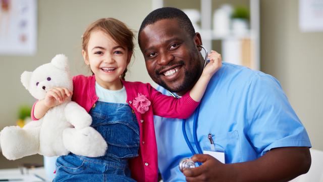 Young child hugs her teddy bear and left arm upright by the healthcare provider's neck both sitting