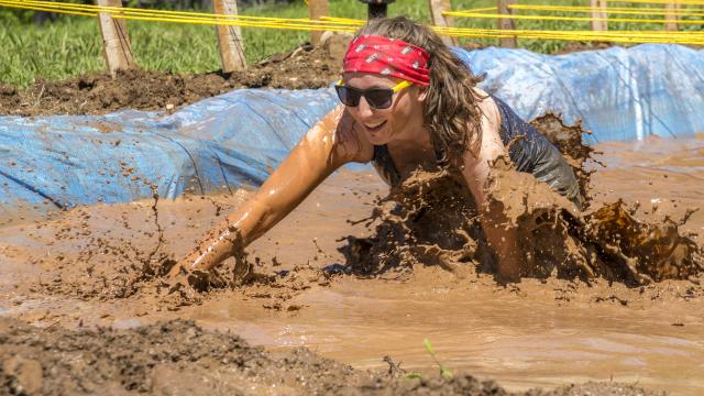 A smiling adult with sunglasses crawling through mud pit with water.