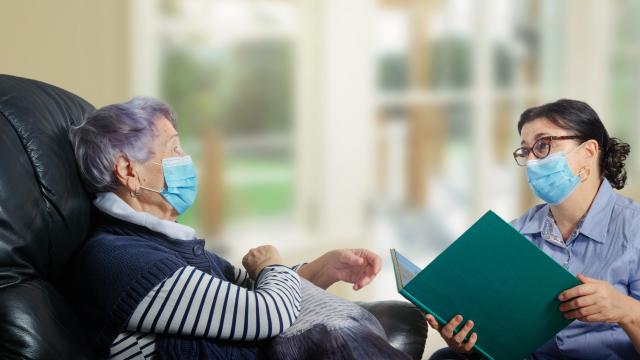 Mask wearing adult reading a book to a mask wearing older adult
