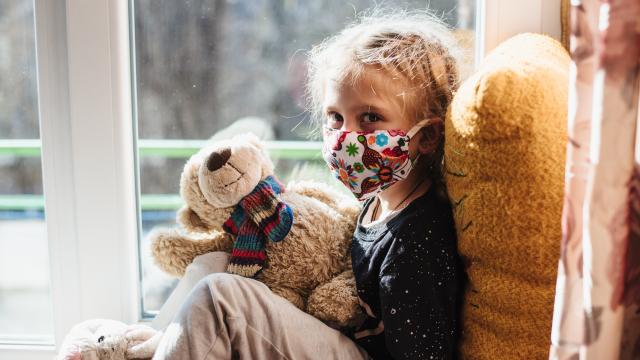 Child wearing a mask and holding a teddy bear sitting in a window seat