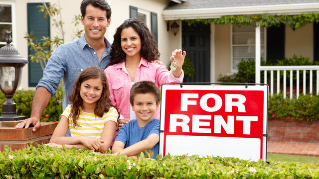 A smiling family stand front of a for rent sign.