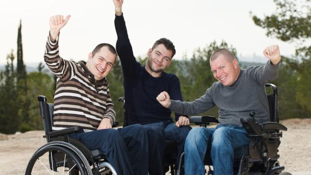 Three people in wheelchairs smile with their thumbs up in the air outside