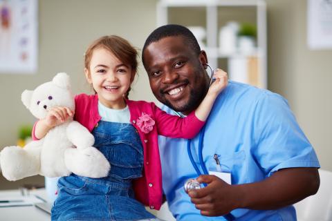 Young child hugs her teddy bear and left arm upright by the healthcare provider's neck both sitting