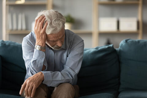 An older adult has trouble remembering while sitting on sofa with his head in his hand.