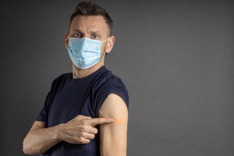A masked adult with sleeve rolled up pointing to his vaccinated arm