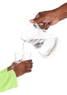 Adult hand pouring water from a pitcher into a glass held by a child