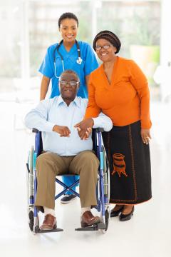 A person in a wheelchair holding hands with another person and a healthcare provider standing behind, all smiling
