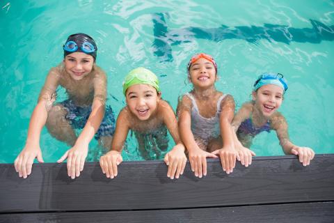 Four young children holding on to the edge of a swimming pool smiling.