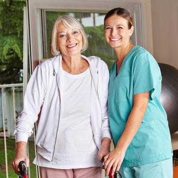 A smiling caregiver standing by a smiling senior with a walker.