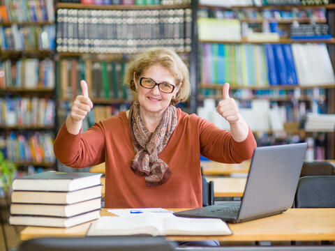 Older adult in a library with books and computer giving two thumbs up,