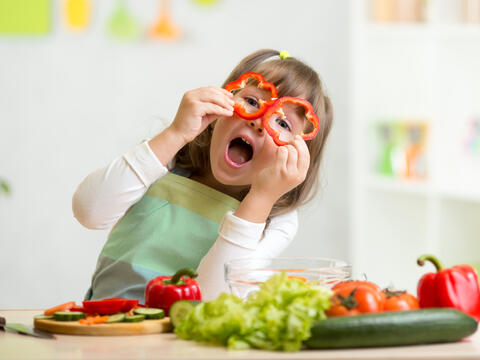 A happy child holding up sliced red peppers as glasses with vegetables and fruits on the table.