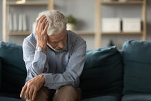 An older adult has trouble remembering while sitting on sofa with his head in his hand.