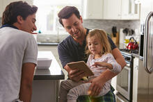 An adult showing a tablet to a child with another adult looking on in a kitchen.