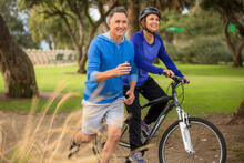 Senior adult runs along an adult riding a bicycle on trail in the park
