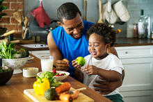 A toddler holding an apple sitting on an adults lap at the kitchen counter next to fruits and vegetables.