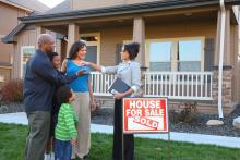 Realtor hands keys to new homeowners with children front of a house with sign for sale saying sold