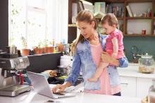An adult holding a toddler checks her laptop while standing in the kitchen